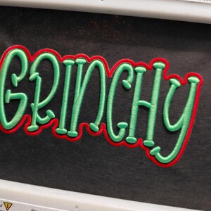 Może przedstawiać: Haftowana naszywka z napisem "GRINCHY" w kolorze zielonym, obramowana na czerwono. Naszywka znajduje się na czarnym tle i jest trzymana w białej tamborku. Na tamborku widnieje napis "8"x13" Mighty Hoop".