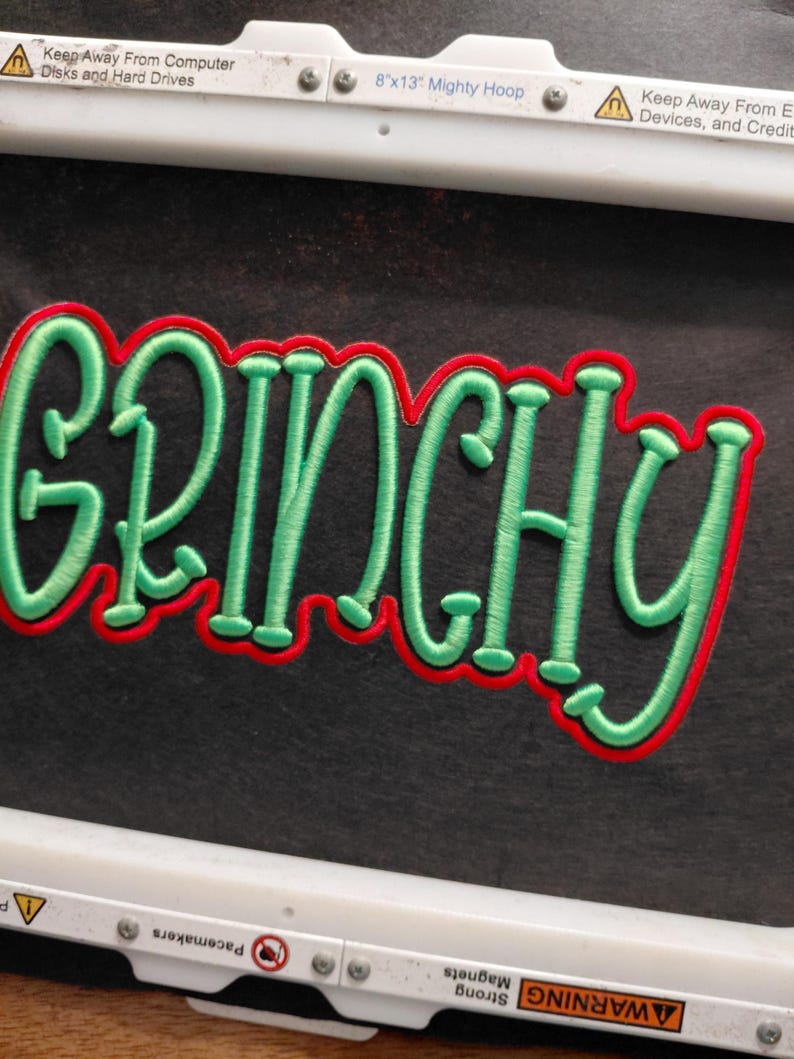Może przedstawiać: Haftowana naszywka z napisem "GRINCHY" w kolorze zielonym, obramowana na czerwono. Naszywka znajduje się na czarnym tle i jest trzymana w białej tamborku. Na tamborku widnieje napis "8"x13" Mighty Hoop".