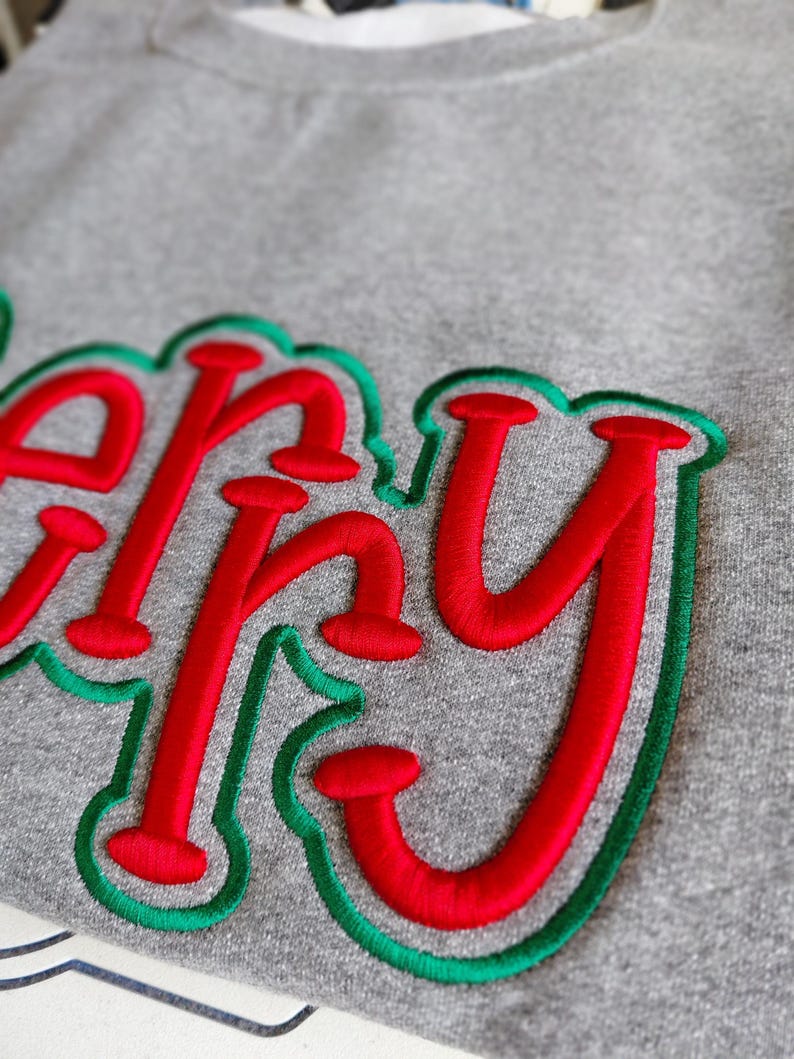 Puede incluir: Primer plano de una sudadera gris con la palabra "Happy" bordada en rojo con un contorno verde. Las letras son grandes y estilizadas, creando un dise&ntilde;o festivo y alegre. La sudadera parece estar hecha de un material suave y c&oacute;modo.
