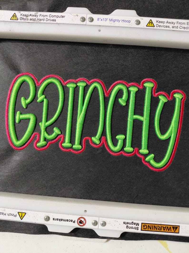 Może przedstawiać: Zielone haftowane słowo "GRINCHY" z czerwonym obrysem na czarnym tle. Słowo jest w zabawnym, kursywnym kroju pisma. Haft jest prawdopodobnie przeznaczony na naszywkę lub aplikację.