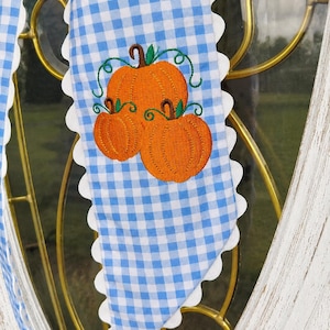 Könnte beinhalten: Ein blau-weiß karierter Stoffstreifen mit weißem Wellenrand, der in der Mitte ein gesticktes Motiv aus drei orangefarbenen Kürbissen mit braunen Stielen und grünen Ranken zeigt. Der Stoff ist über einen goldfarbenen Metallrahmen drapiert.