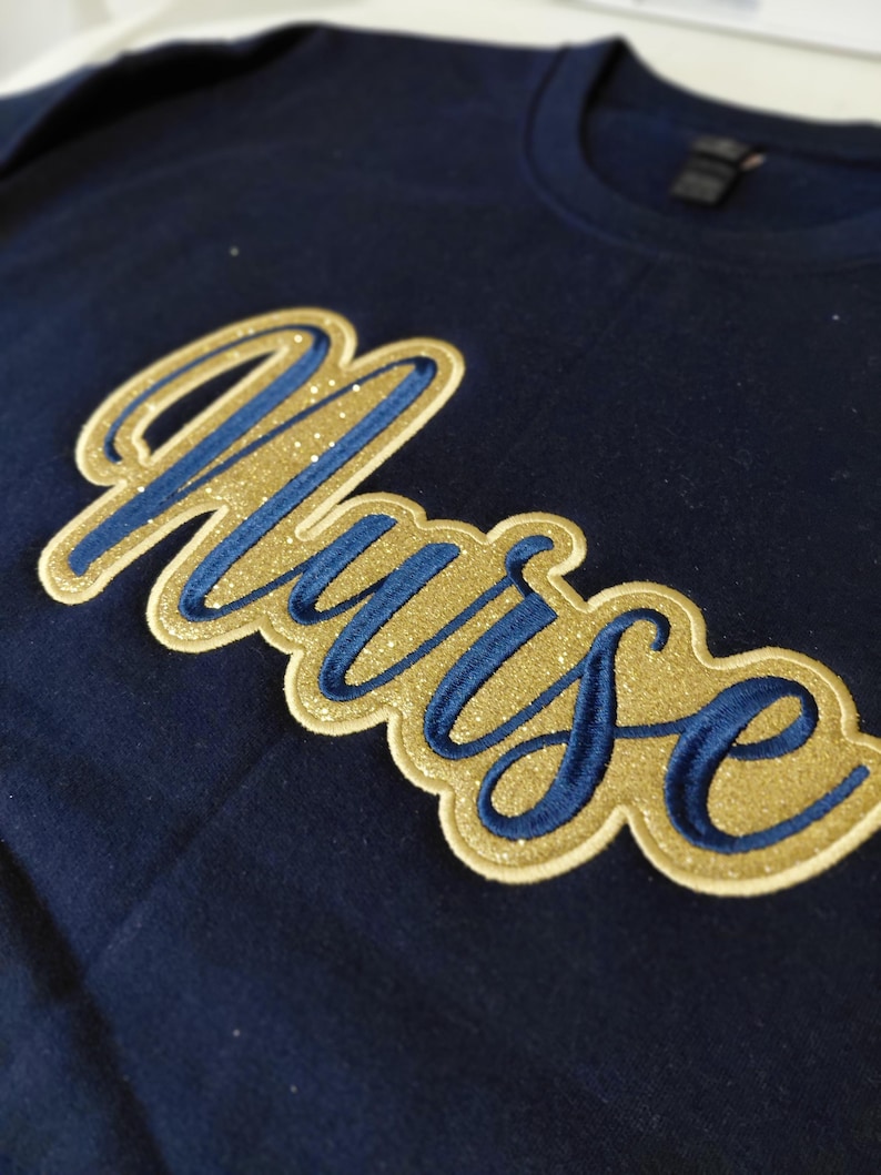 May include: Navy blue t-shirt with the word "Nurse" embroidered in gold glitter and blue thread. The word is in a cursive font and has a gold border. The shirt is made of a soft material.