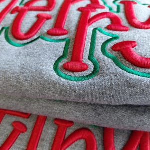 Puede incluir: Primer plano de una sudadera gris con la palabra "Merry" bordada en hilo rojo, contorneada en verde. La sudadera est&aacute; doblada, mostrando el texto bordado en dos capas. La tela tiene un aspecto suave y texturizado.