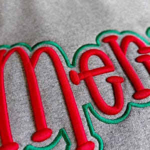 Puede incluir: Primer plano de una sudadera gris con la palabra "merry" bordada en rojo con un contorno verde. El bordado tiene un aspecto texturizado y en relieve. La sudadera est&aacute; hecha de un material suave y c&oacute;modo.