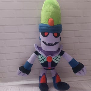 May include: A plush doll of a cartoon character with a green hat, blue headband, and purple skin. The doll has red eyes, a black mouth, and a black and purple outfit with red and blue accents. The doll is standing in front of a white brick wall.