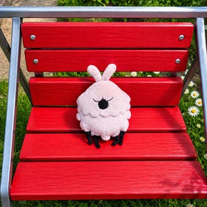 May include: A plush, light pink creature with a round body, small black legs, and two upright ears, resting on a red bench. The creature has a black, closed-eye design. The bench is made of red painted wood and has a metal frame.