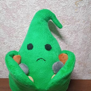 Puede incluir: Un peluche verde con forma de criatura con cabeza puntiaguda, ojos negros y boca curvada. La criatura sostiene varias piedras pequeñas, coloridas y redondeadas. El juguete está hecho de tela suave y tiene un diseño caprichoso.