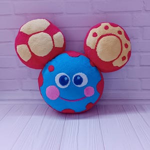 May include: A soft toy featuring a smiling face with three circular elements. The central face is blue with pink cheeks and red spots. Two red and yellow circles sit above, like ears. The toy rests on a white and pink surface.