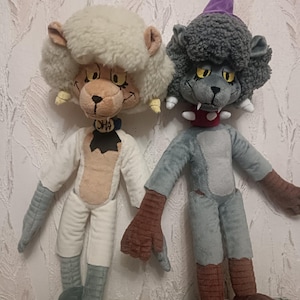 Puede incluir: Dos muñecos de peluche con rasgos de león de dibujos animados. Uno tiene una melena blanca y esponjosa y un atuendo blanco, el otro tiene una melena y un atuendo grises, y un sombrero morado. Ambos tienen manos y pies grandes, y están sobre una superficie verde estampada.