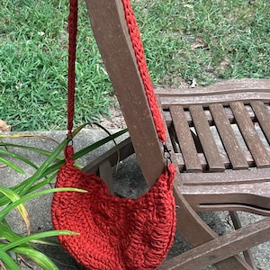 May include: A handmade, red crochet shoulder bag with a long strap, hanging on a brown wooden chair. The bag has a crescent shape and a textured surface, perfect for carrying essentials. The background features green grass and foliage.