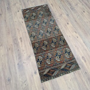 Puede incluir: Una alfombra rectangular con un patrón geométrico repetido en tonos marrón, azul y crema. El borde de la alfombra presenta formas de diamante. Se coloca sobre un suelo de madera clara. Las dimensiones de la alfombra no son visibles.