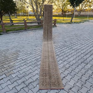 Op de afbeelding: Een lange, rechthoekige loper met een gedetailleerd, herhalend patroon in tinten beige en bruin. De loper wordt buiten op een bakstenen oppervlak getoond, waarbij de lengte en het ingewikkelde ontwerp worden getoond. De randen van de loper zijn afgewerkt met een rode rand.
