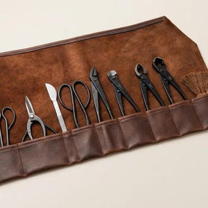 Op de afbeelding: Een bruine lederen gereedschapset met een set zwarte metalen bonsaigereedschappen, waaronder diverse scharen, tangen, snijders, een klein mes en een houten borstel, georganiseerd in individuele gestikte vakken in de open lederen hoes.