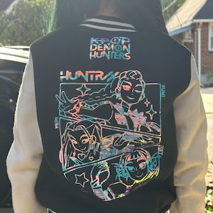 May include: Black varsity jacket with white sleeves. The back features a colorful graphic of anime-style characters and the text "K-POP DEMON HUNTERS" and "HUNTRX". The jacket has a striped collar and cuffs.