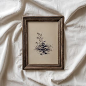 Pode incluir: Ilustra&ccedil;&atilde;o bot&acirc;nica a preto e branco emoldurada de uma cobra enrolada em plantas e flores. A obra de arte est&aacute; numa moldura de madeira castanha escura, sobre um fundo de tecido branco.
