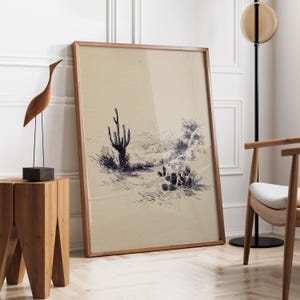 May include: A framed desert landscape art print featuring a pen and ink drawing of cacti and desert plants. The artwork is in a wooden frame and leans against a white wall. A wooden sculpture of a bird and a side table are in the foreground.