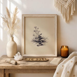 Pode incluir: Uma impress&atilde;o emoldurada em estilo vintage de uma cobra enrolada perto de flores e grama. A impress&atilde;o est&aacute; em uma moldura de madeira de cor clara. A imagem est&aacute; em uma mesa de madeira com um vaso de flores secas, um cristal, um bast&atilde;o de fuma&ccedil;a, uma vela e uma bandeja de fases da lua.