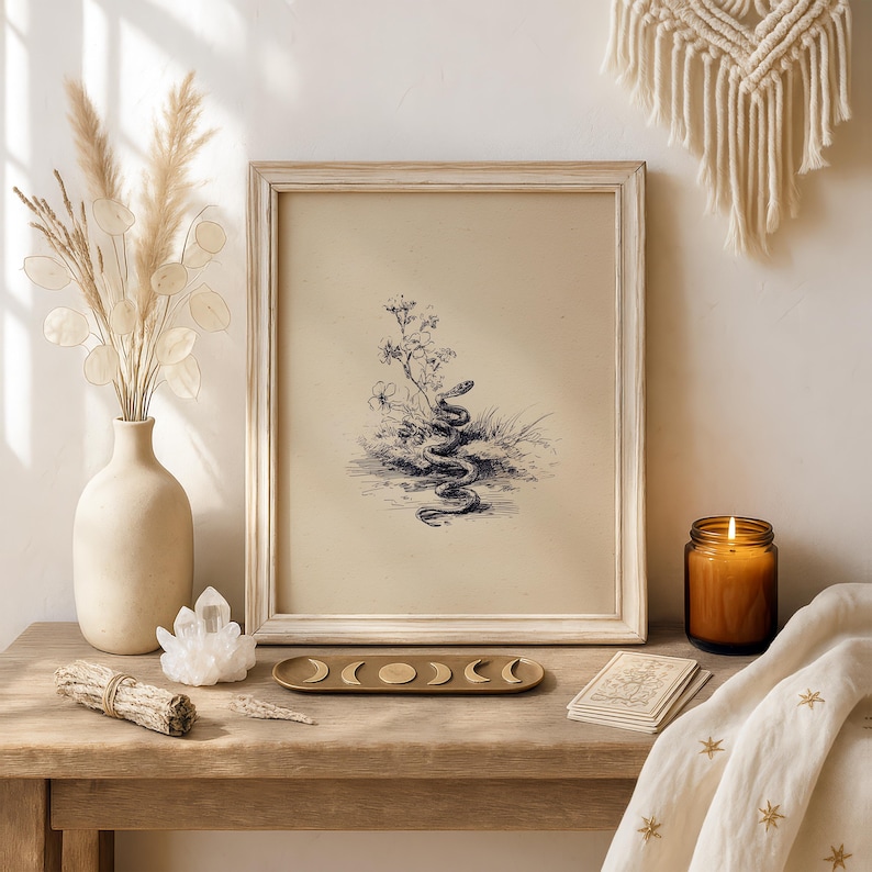 Pode incluir: Uma impress&atilde;o emoldurada em estilo vintage de uma cobra enrolada perto de flores e grama. A impress&atilde;o est&aacute; em uma moldura de madeira de cor clara. A imagem est&aacute; em uma mesa de madeira com um vaso de flores secas, um cristal, um bast&atilde;o de fuma&ccedil;a, uma vela e uma bandeja de fases da lua.