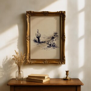 May include: A framed desert landscape art print with a gold ornate frame. The artwork features a blue and white sketch of a cactus and other desert plants. The frame is hanging above a wooden table with books, a vase, and a candle holder.