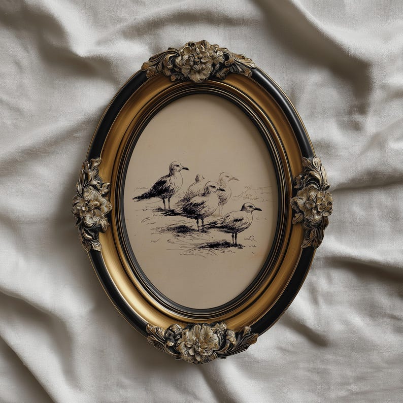 May include: Oval picture frame with a black and gold finish, featuring a pen and ink drawing of seagulls. The frame has ornate floral accents at the top and bottom. The artwork is set against a cream-colored background.