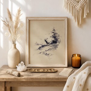 May include: A framed vintage-style print of a deer on a hillside, in a light-colored wooden frame. The image is in black and white. The scene is set on a wooden table with a vase of dried flowers, a candle, and decorative items.