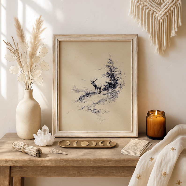 May include: A framed vintage-style print of a deer on a hillside, in a light-colored wooden frame. The image is in black and white. The scene is set on a wooden table with a vase of dried flowers, a candle, and decorative items.