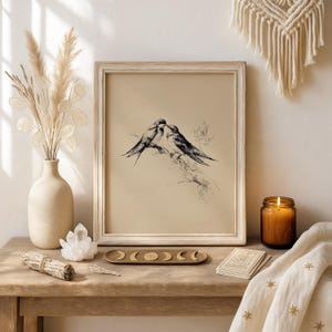 May include: A framed print featuring a black and white illustration of two birds perched on a branch. The artwork is displayed on a wooden table, alongside a vase with dried flowers, a candle, and decorative items. The frame is a light, neutral color.