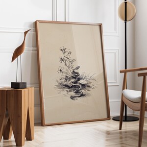Pode incluir: Uma impress&atilde;o emoldurada com uma ilustra&ccedil;&atilde;o em preto e branco de uma cobra em meio a flores e grama. A obra de arte tem uma est&eacute;tica vintage, com um fundo bege claro e uma moldura de madeira. A impress&atilde;o est&aacute; encostada em uma parede branca.
