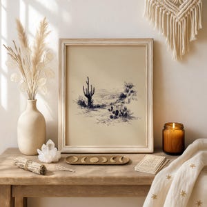 May include: A framed black and white desert landscape art print featuring a cactus. The print is in a light-colored wooden frame. The image is on a wooden table with a vase of dried flowers, a candle, and other decorative items.