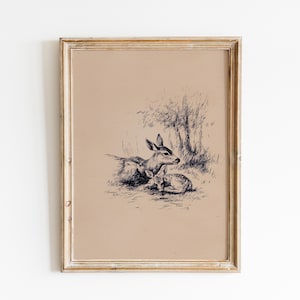 May include: A framed pen and ink drawing of two deer resting in a grassy area. The artwork features a mother deer and a fawn, rendered in black and white, set against a tan background. The frame is a light, distressed gold color.