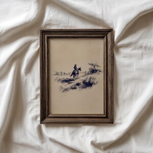 K&ouml;nnte beinhalten: Gerahmte Federzeichnung eines Cowboys auf einem Pferd, vor einer Landschaft mit einem Zaun und sp&auml;rlicher Vegetation. Das Kunstwerk befindet sich in einem braunen Holzrahmen und erzeugt eine Vintage-&Auml;sthetik.