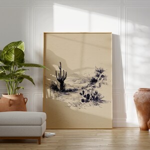 May include: A framed print featuring a desert landscape sketch in black ink on a beige background. The artwork depicts a cactus, other desert plants, and distant hills. The print is displayed in a room with a plant, a footstool, and a terracotta pot.