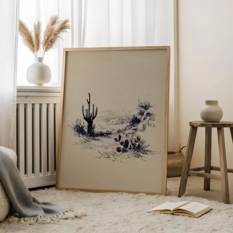 May include: A framed print featuring a desert landscape with a cactus and other desert plants. The artwork is rendered in a pen and ink style with blue ink on a cream-colored background. The frame is made of light-colored wood.