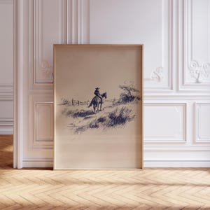 K&ouml;nnte beinhalten: Gerahmte Tuschezeichnung, die einen Cowboy auf einem Pferd in einer grasbewachsenen Landschaft zeigt. Das Kunstwerk, in Braun- und Marineblaut&ouml;nen, zeigt einen Zaun und sp&auml;rliche Vegetation unter einem klaren Himmel. Die gerahmte Kunst wird vor einer wei&szlig;en Wand ausgestellt.