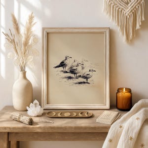May include: A framed print featuring a pen and ink drawing of five seagulls. The artwork is displayed on a wooden table with a vase of dried flowers, a candle in a glass jar, and a decorative tray with moon phase designs.