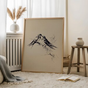 May include: A framed print featuring a black and white illustration of two birds perched on a branch. The artwork has a vintage aesthetic, with a neutral background and a light wooden frame. The print is displayed in a cozy, well-lit room.