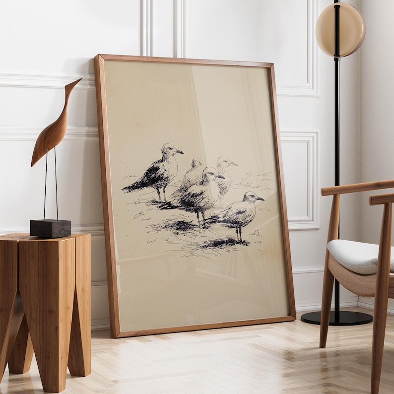 May include: A framed print featuring a pen and ink drawing of several seagulls. The artwork is in a wooden frame and leans against a white wall. A wooden sculpture of a bird and a wooden side table are in the foreground. The floor is made of light-colored wood.