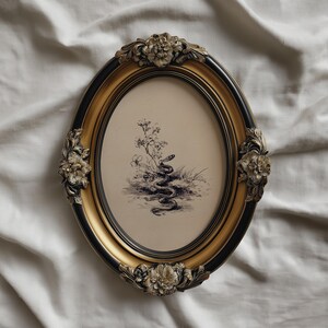 Pode incluir: Uma moldura oval com acabamento preto e dourado. A moldura apresenta enfeites florais e cont&eacute;m uma ilustra&ccedil;&atilde;o a preto e branco de uma cobra num cen&aacute;rio natural. A moldura tem aproximadamente 25 cm de altura.