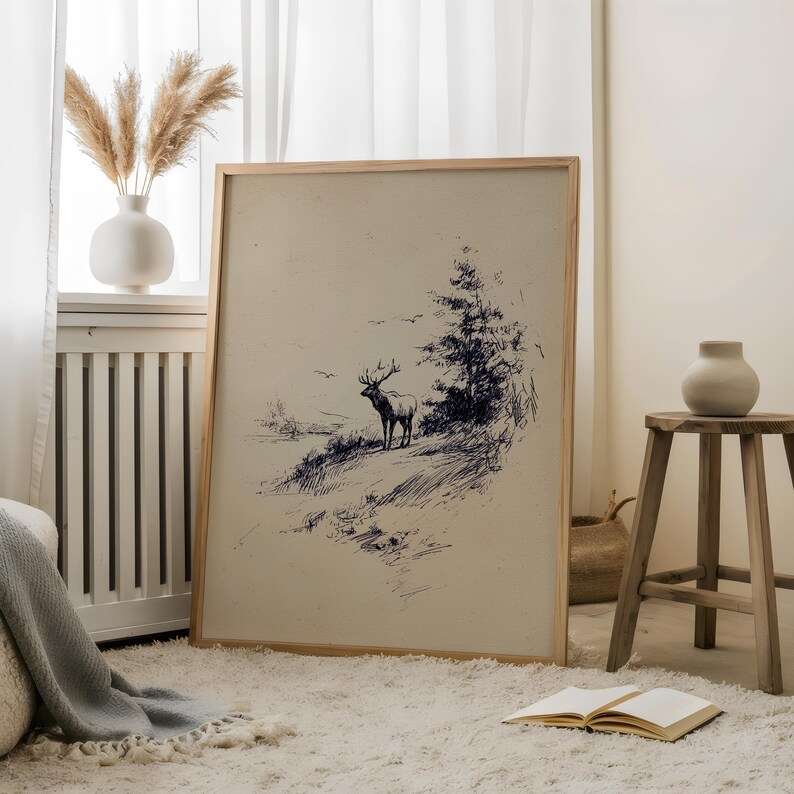 May include: A framed art print featuring a pen and ink drawing of a deer in a natural landscape. The artwork is in a wooden frame and rests on a white rug. A vase with dried flowers and a small wooden stool are also in the room.