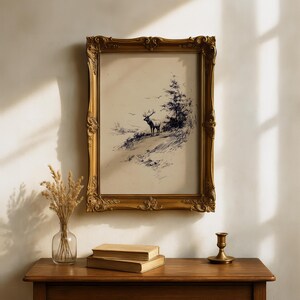 May include: A framed vintage print featuring a detailed pen and ink drawing of a deer standing on a hillside, with trees in the background. The ornate gold frame adds a classic touch to the artwork. The print is displayed above a wooden table.