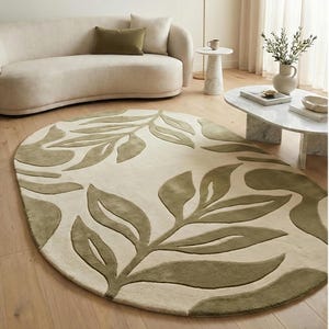 May include: An oval area rug with a cream background and a large olive green leaf design. The rug is placed on a light wood floor, and a beige sofa and marble coffee table are in the background.