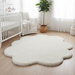 May include: A fluffy, white cloud-shaped rug sits on a light wood floor in a nursery. The rug is the focal point, with a soft, textured surface. A white crib, chair, and plant in a woven basket are in the background, creating a serene atmosphere.
