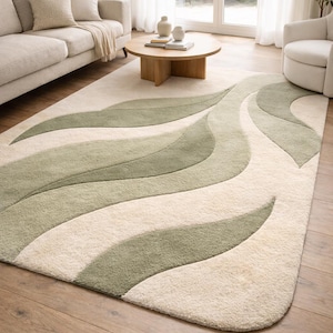 May include: A large, rectangular area rug with a cream-colored background and flowing, leaf-like patterns in shades of green. The rug is placed on a hardwood floor in a living room setting, with a sofa and coffee table visible.