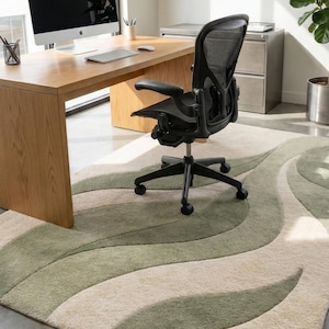 May include: An office scene with a wooden desk, a black mesh office chair, and a large area rug. The rug features a wavy pattern in shades of green and beige. A computer monitor and whiteboard are also present.