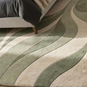 May include: A patterned rug with a flowing, wave-like design. The rug features alternating stripes of light green, medium green, and off-white. The rug is placed near a bed with a grey duvet cover and a beige throw.