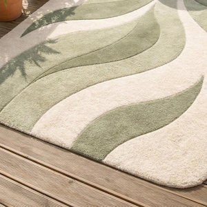 May include: A plush rug with a flowing, organic design. The rug features alternating bands of cream and shades of green, creating a wave-like pattern. The rug is placed on a wooden deck, adding a touch of natural elegance to the space.