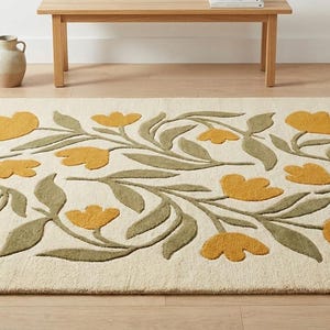 May include: A rectangular rug with a cream-colored background and a floral design. The flowers are mustard yellow, and the leaves are olive green. The rug is placed on a wooden floor, with a wooden bench and a vase in the background.