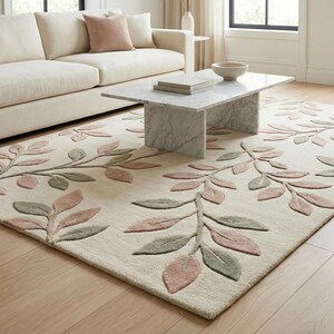 May include: A rectangular area rug with a cream background and a repeating leaf pattern in shades of pink and green. The rug is placed on a hardwood floor in a living room setting, with a beige sofa and marble coffee table.