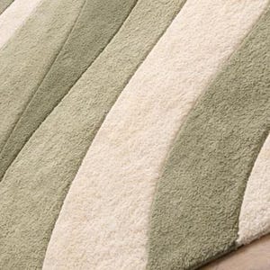 May include: Close-up of a soft rug with a wave design. The rug has alternating stripes of light green, off-white, and medium green. The rug is made of a plush material, creating a cosy and inviting feel. The rug is likely made of synthetic fibres.