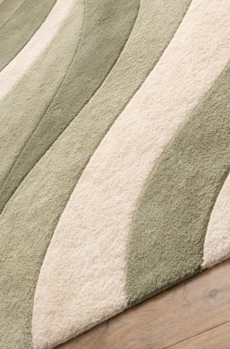 May include: Close-up of a soft rug with a wave design. The rug has alternating stripes of light green, off-white, and medium green. The rug is made of a plush material, creating a cosy and inviting feel. The rug is likely made of synthetic fibres.